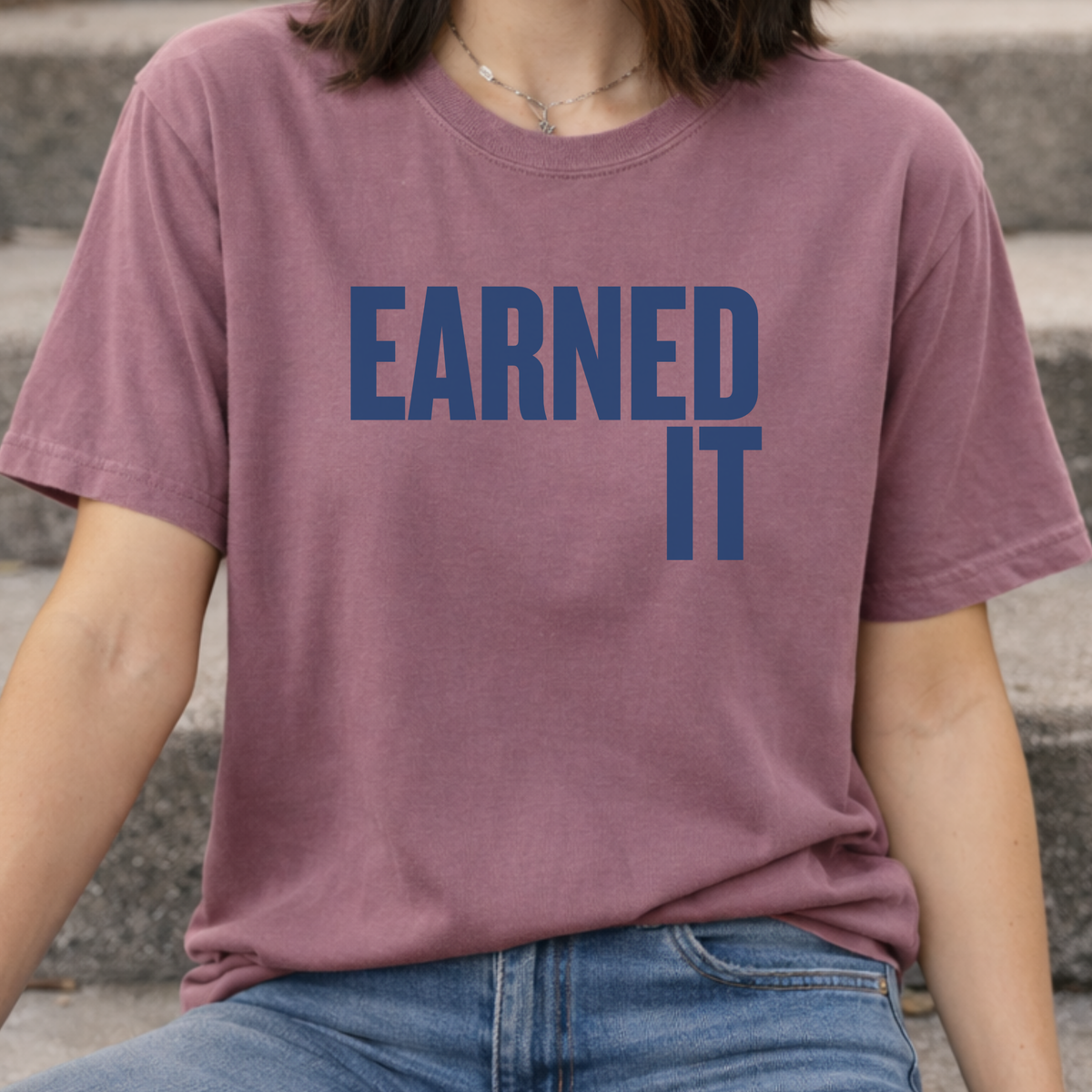 A woman in a berry colored Comfort Colors t-shirt that says "Earned It" in navy blue.
