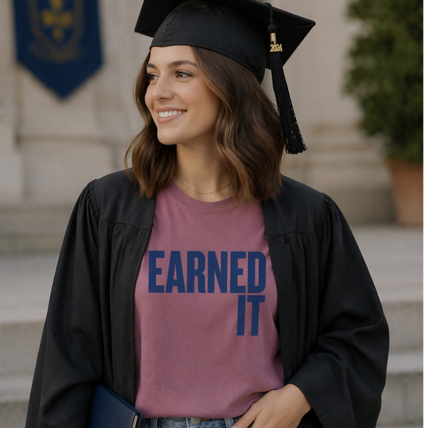 A woman with a graduation cap and gown on with a t-shirt that says "Earned It" in navy blue.