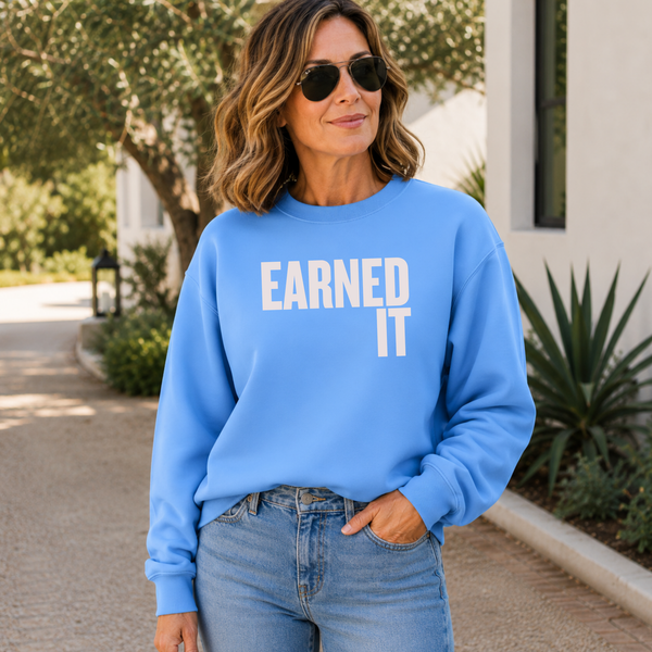 A woman standing outside in a light blue sweatshirt with the words "Earned It" in light pink font.