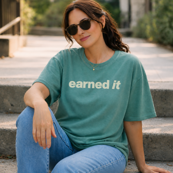 A woman in jeans and a light green tee shirt that says "earned it" in beige sitting on the stairs.