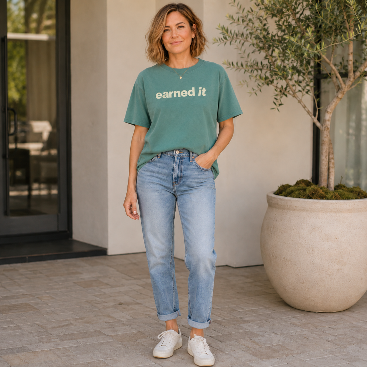 A woman in jeans and a light green tee shirt that says "earned it" in beige standing outside in jeans and sneakers