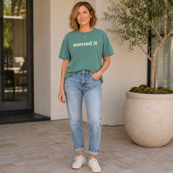 A woman in jeans and a light green tee shirt that says "earned it" in beige standing outside in jeans and sneakers