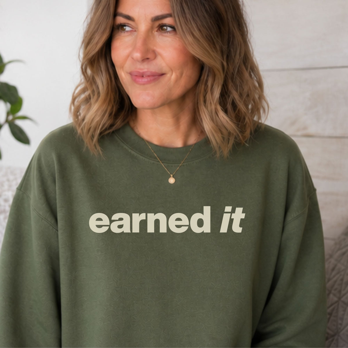 A woman sitting in a dark green sweatshirt with the words "Earned It" in a soft celery yellow font.