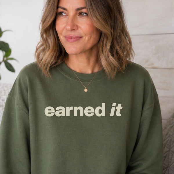 A woman sitting in a dark green sweatshirt with the words "Earned It" in a soft celery yellow font.