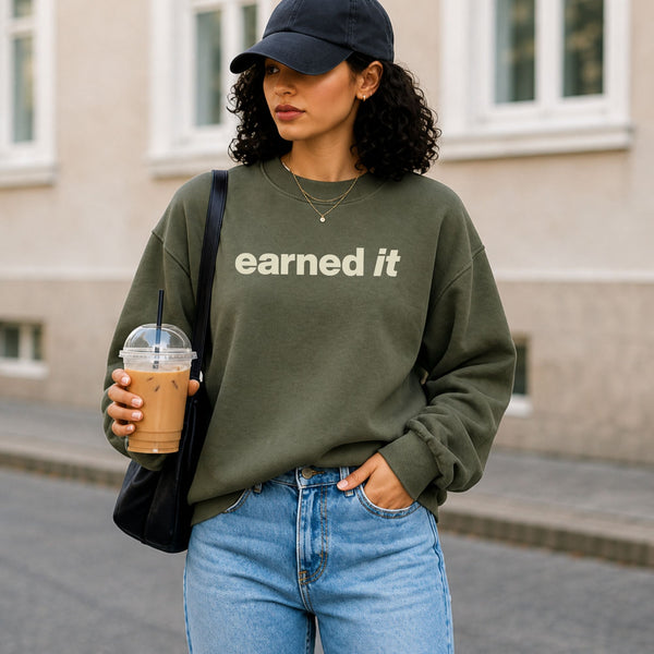 A woman outside in a dark green sweatshirt with the words "Earned It" in a soft celery yellow font, holding an iced coffee.