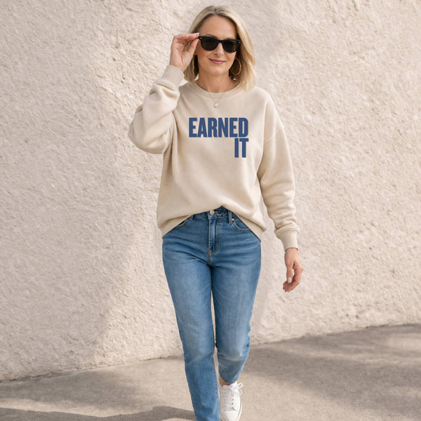 A woman walking outdoors wearing a beige sweatshirt and jeans. The sweatshirt says "Earned It" in navy font.