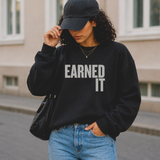 Woman wearing a black sweatshirt with 'EARNED IT' text, blue jeans, and a black cap outdoors.