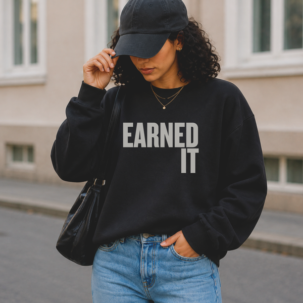 Woman wearing a black sweatshirt with 'EARNED IT' text, blue jeans, and a black cap outdoors.