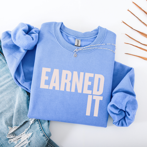 A folded light blue sweatshirt with the words "Earned It" in soft pink font.