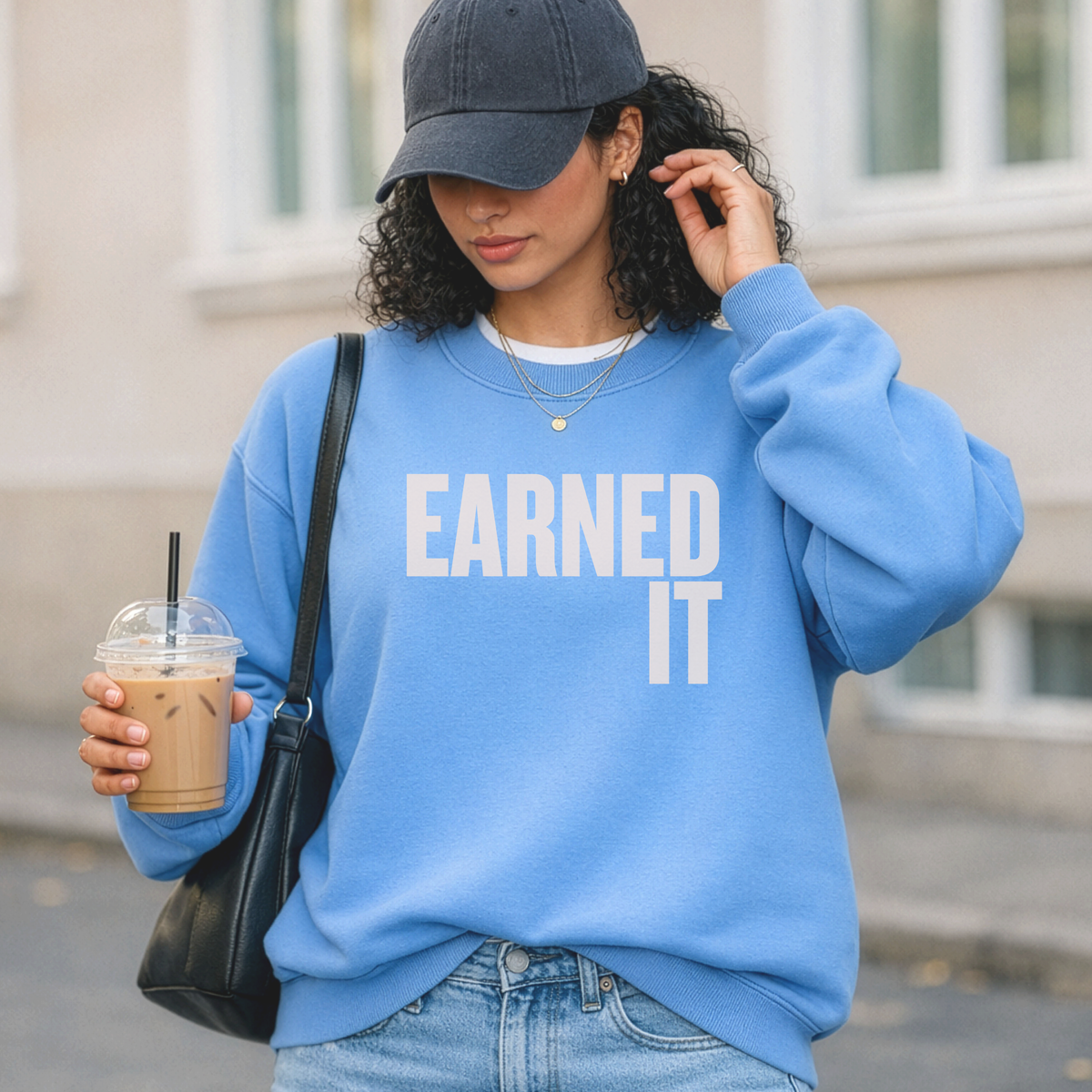 A woman standing outside in a light blue sweatshirt with the words "Earned It" in light pink font holding an iced coffee.
