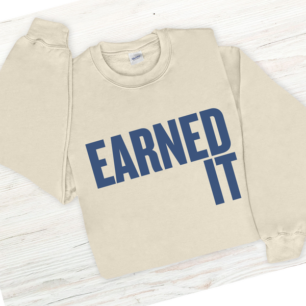 Beige sweatshirt with 'Earned It' text in blue on a wooden surface