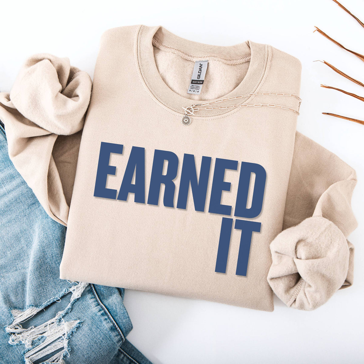 A folded beige sweatshirt and jeans. The sweatshirt says "Earned It" in navy font.