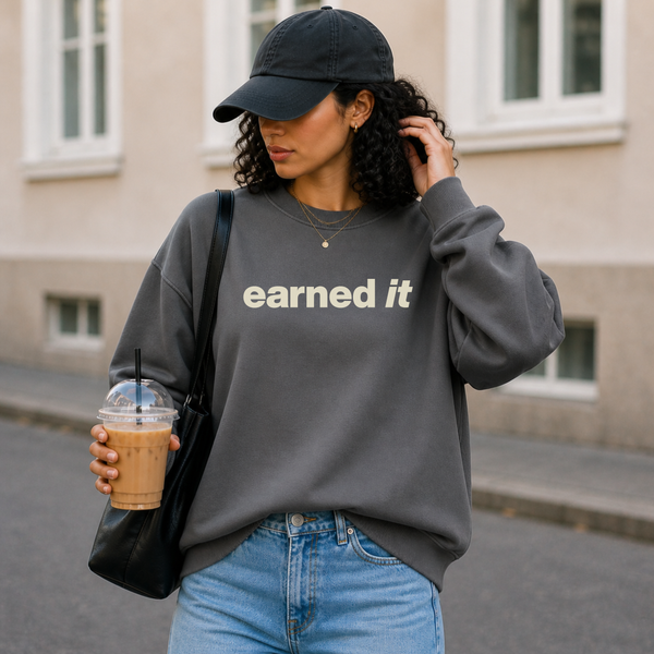 A woman outside in a charcoal gray sweatshirt with the words "Earned It" in a soft celery yellow font.