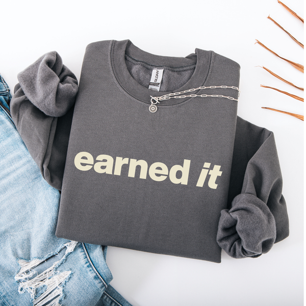 A dark gray folded sweatshirt with the words "Earned It" in a soft celery yellow font.
