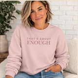 A woman wearing a light pink crewneck sweatshirt with the text 'THAT'S ABOUT ENOUGH' printed in a dusty rose font across the chest, paired with blue jeans in a cozy indoor setting.