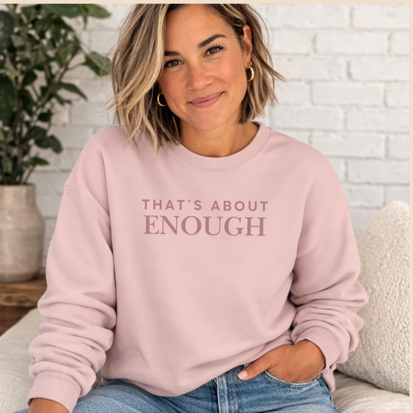 A woman wearing a light pink crewneck sweatshirt with the text 'THAT'S ABOUT ENOUGH' printed in a dusty rose font across the chest, paired with blue jeans in a cozy indoor setting.