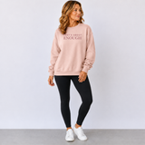 A woman modeling a light pink crewneck sweatshirt with the text 'WHAT'S ABOUT ENOUGH' printed in dark pink across the chest, paired with black leggings and white sneakers against a light gray studio background.