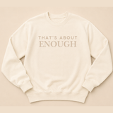 An off-white crewneck sweatshirt with the text 'THAT'S ABOUT ENOUGH' printed in a muted taupe-brown serif font across the chest, displayed flat against a matching warm beige background.