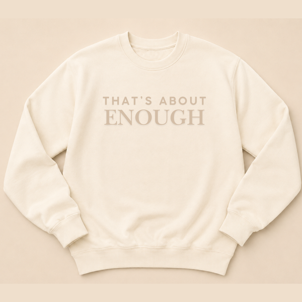 An off-white crewneck sweatshirt with the text 'THAT'S ABOUT ENOUGH' printed in a muted taupe-brown serif font across the chest, displayed flat against a matching warm beige background.
