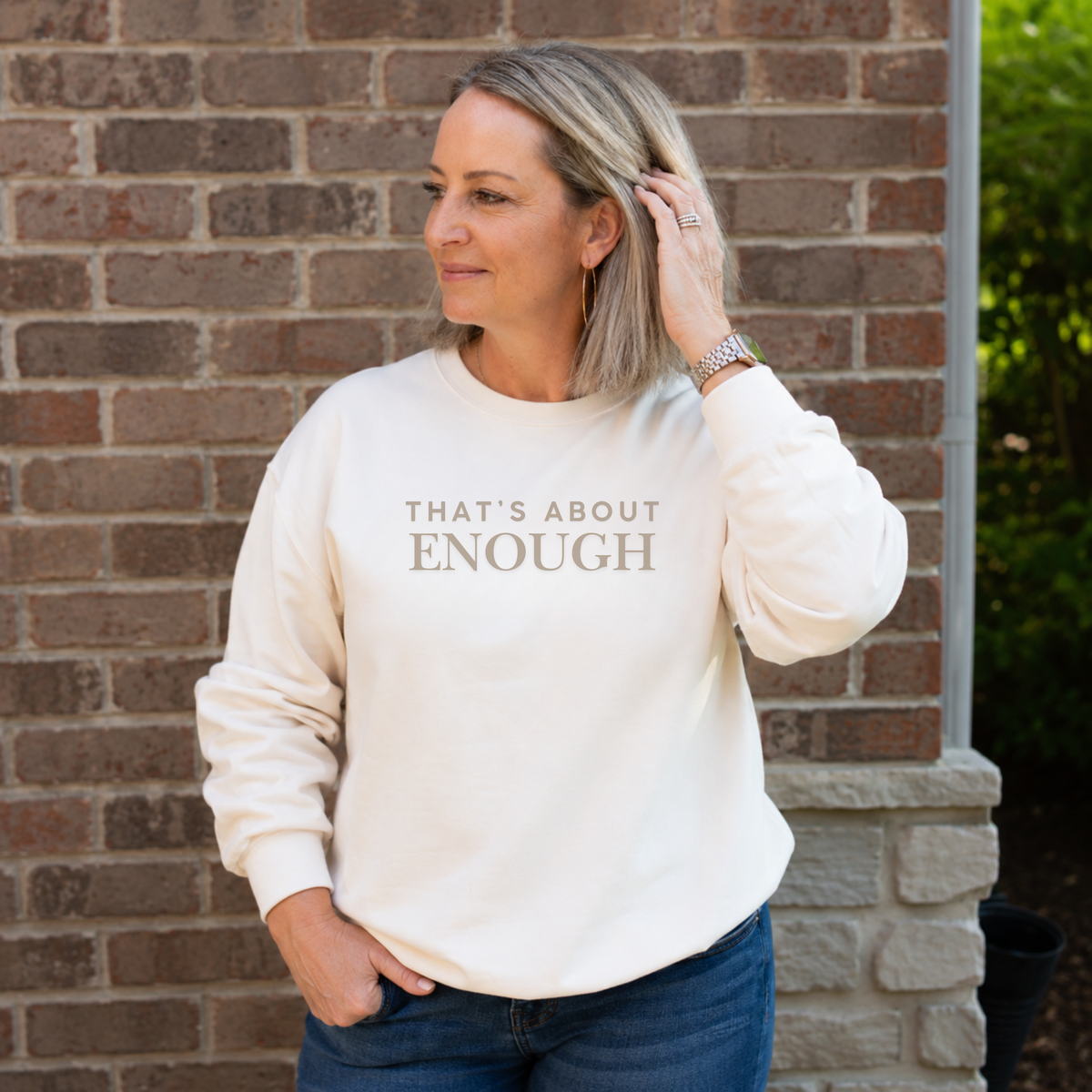 A woman wearing a cream-colored crewneck sweatshirt with the text 'THAT'S ABOUT ENOUGH' printed in gray on the front, standing in front of a brick wall.