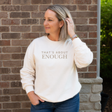 A woman wearing a cream-colored crewneck sweatshirt with the text 'THAT'S ABOUT ENOUGH' printed in gray on the front, standing in front of a brick wall.