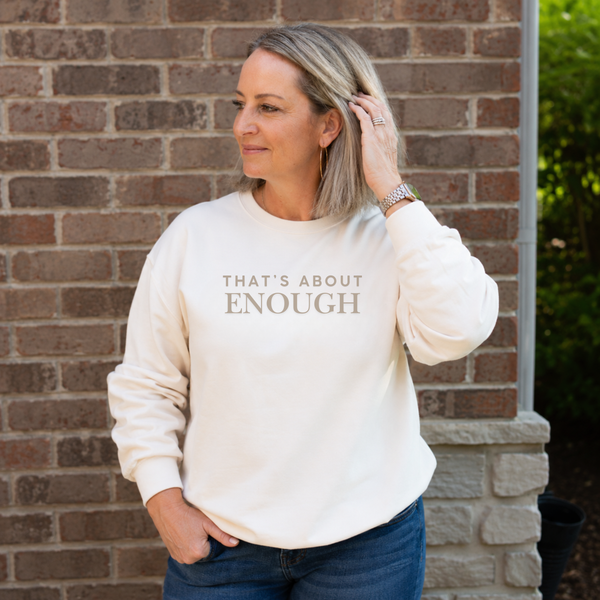 A woman wearing a cream-colored crewneck sweatshirt with the text 'THAT'S ABOUT ENOUGH' printed in gray on the front, standing in front of a brick wall.