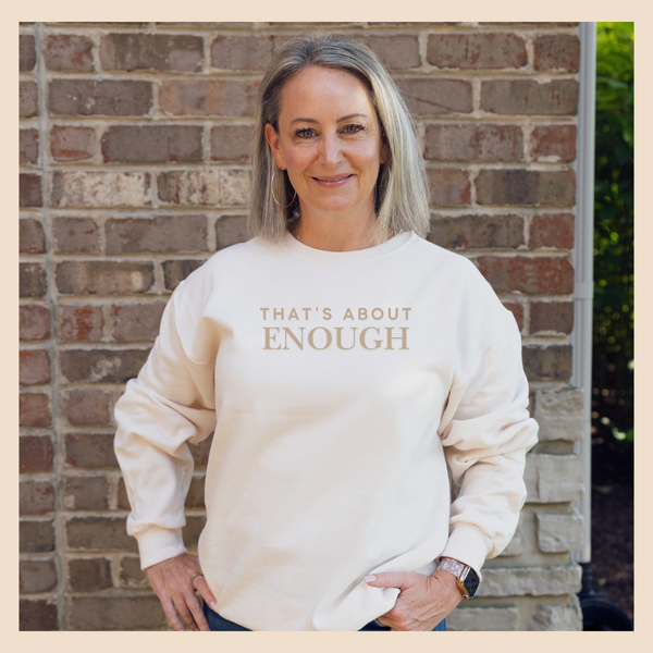 A woman with gray hair stands in front of a brick wall wearing a cream-colored sweatshirt with the text 'THAT'S ABOUT ENOUGH' printed in a metallic gold tone across the chest.