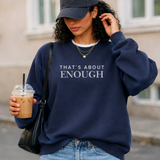 A woman wearing a navy blue sweatshirt with the text 'THAT'S ABOUT ENOUGH' in white, styled with a navy baseball cap, light blue jeans, a black shoulder bag, and an iced coffee in an outdoor urban setting.