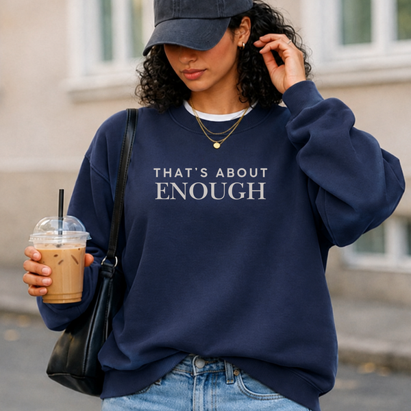 A woman wearing a navy blue sweatshirt with the text 'THAT'S ABOUT ENOUGH' in white, styled with a navy baseball cap, light blue jeans, a black shoulder bag, and an iced coffee in an outdoor urban setting.