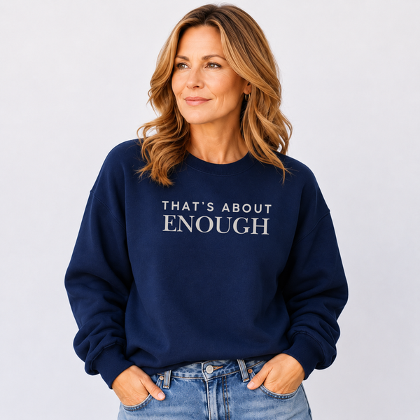 A woman wearing a navy blue crewneck sweatshirt with the text 'THAT'S ABOUT ENOUGH' printed in white across the chest, paired with light-wash blue jeans, set against a plain light gray background.