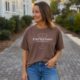 Woman wearing a brown t-shirt with 'Espresso Yourself' text and a glass graphic, standing on a paved path.