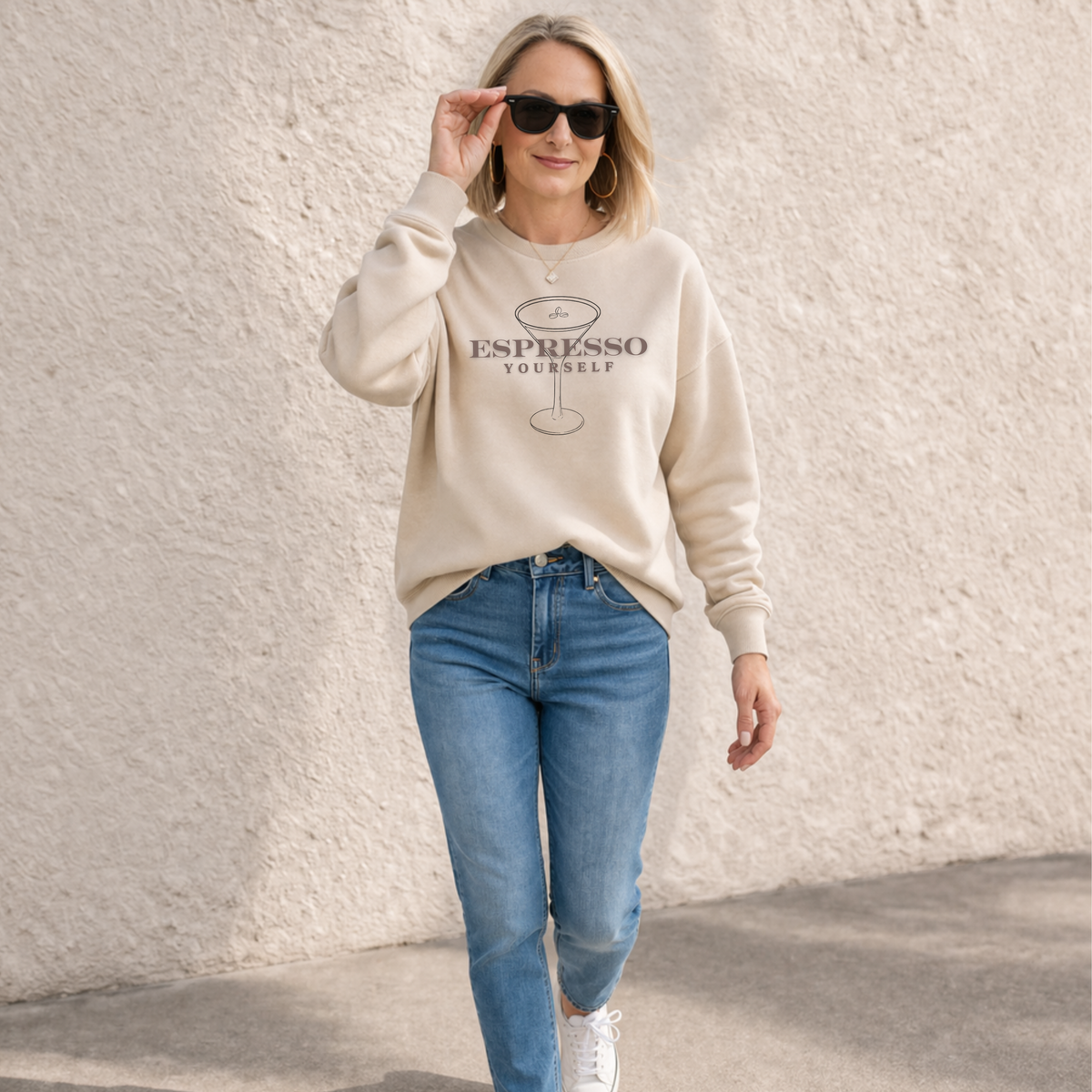 Woman wearing a beige sweatshirt with text and graphics, blue jeans, and sunglasses against a light textured wall.