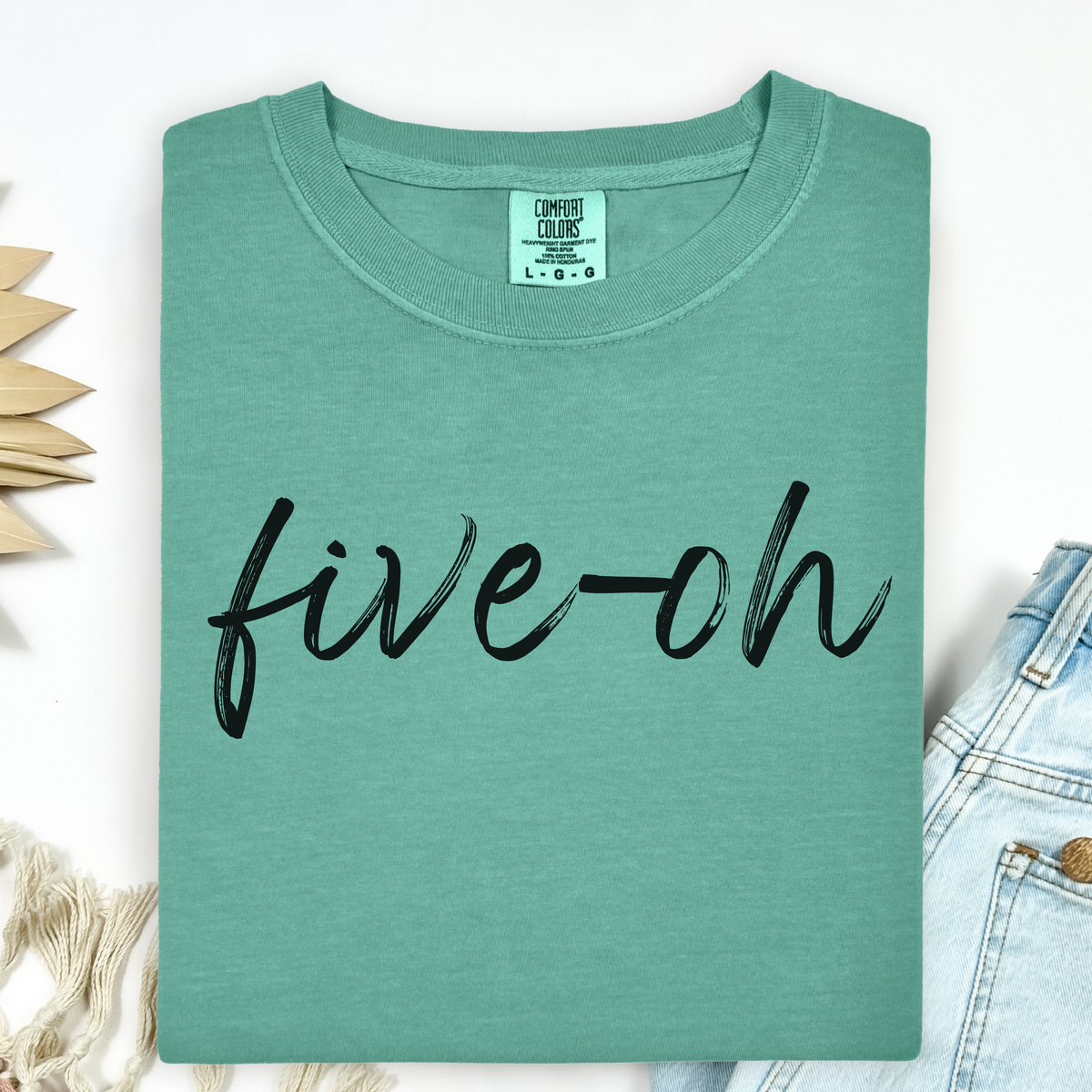 A folded Comfort Colors Green t-shirt with 'five-oh' text on a white background, sitting next to a pair of jeans.