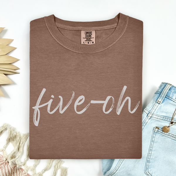 A folded Comfort Colors Espresso t-shirt with 'five-oh' text on a white background, sitting next to a pair of jeans.