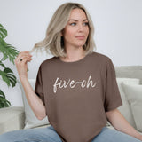 Woman wearing a brown t-shirt with 'five-oh' text, sitting on a couch.