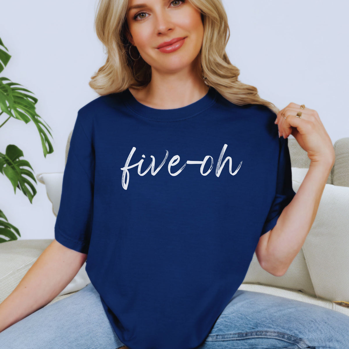 Woman wearing jeans and a navy blue t-shirt with 'five-oh' text, sitting on a couch.