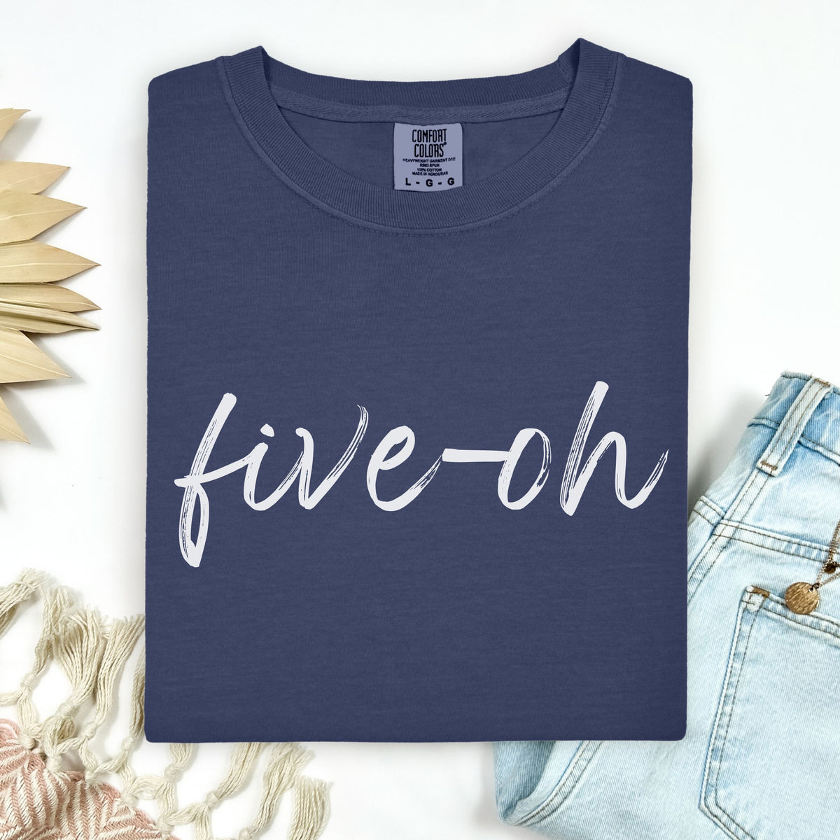 A folded Comfort Colors Navy blue t-shirt with 'five-oh' text on a white background, sitting next to a pair of jeans.