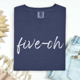 A folded Comfort Colors Navy blue t-shirt with 'five-oh' text on a white background, sitting next to a pair of jeans.