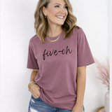 Woman wearing a mauve t-shirt with 'five-oh' text, standing against a white wall.