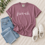 A mauve short-sleeve T-shirt with the phrase 'five-oh' printed in white script across the chest, styled in a flat lay with light blue jeans, white sneakers, and green eucalyptus leaves on a cream fabric background.