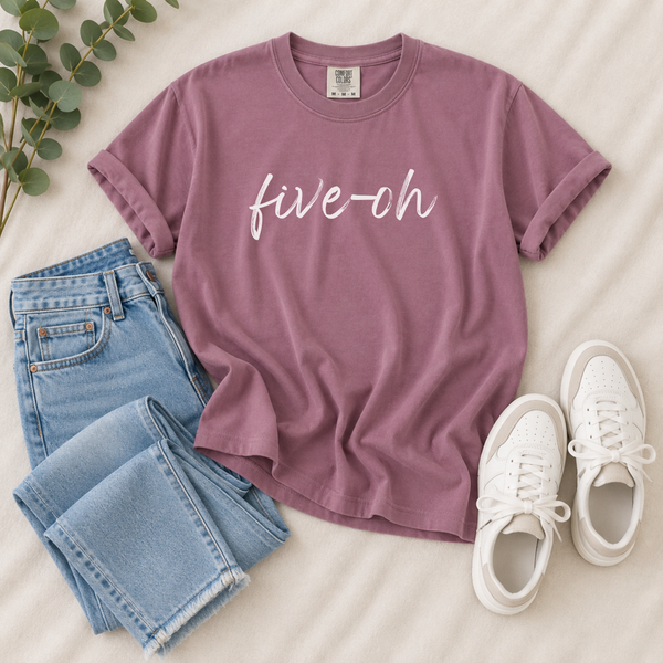 A mauve short-sleeve T-shirt with the phrase 'five-oh' printed in white script across the chest, styled in a flat lay with light blue jeans, white sneakers, and green eucalyptus leaves on a cream fabric background.