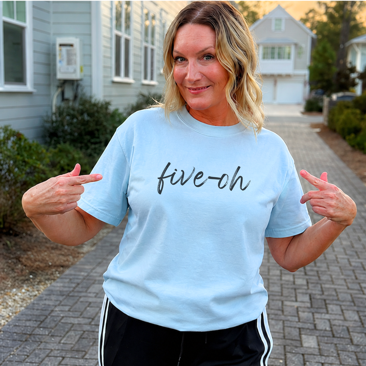A woman wearing a light blue graphic t-shirt with the text 'five-ch' in black script, posing on a paved walkway in front of houses.
