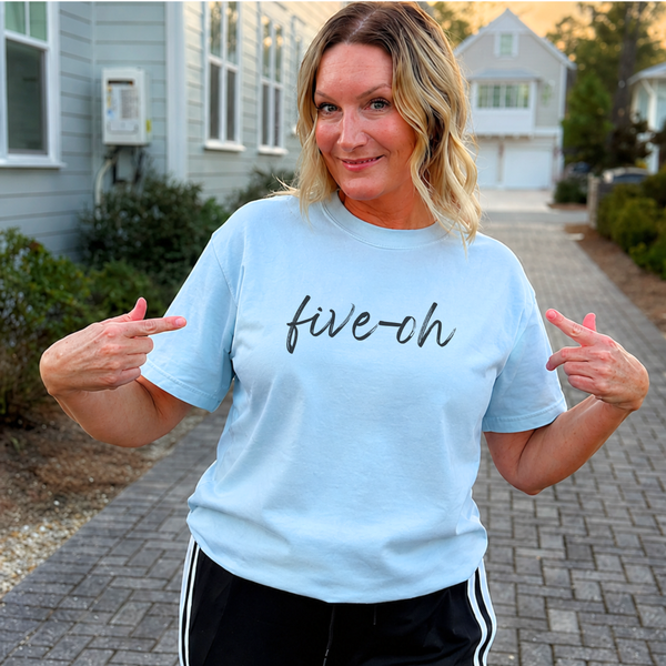 A woman wearing a light blue graphic t-shirt with the text 'five-ch' in black script, posing on a paved walkway in front of houses.