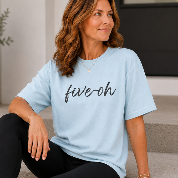 A woman sits on concrete steps wearing a light blue oversized T-shirt with the words 'five-oh' printed across the chest in black script, paired with black leggings.