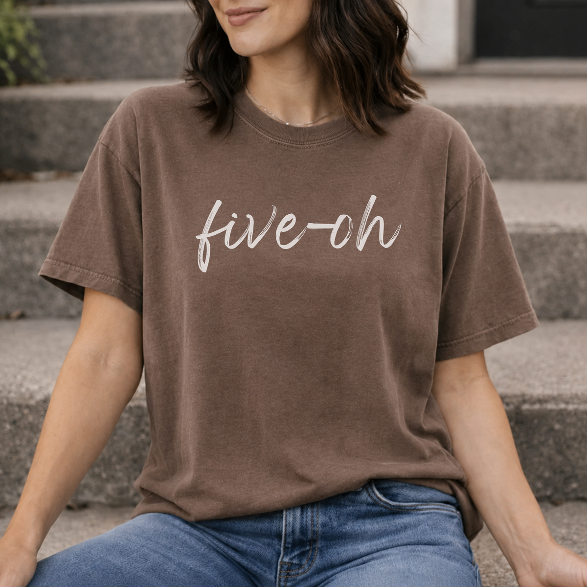 A woman sitting indoors with a espresso brown colored Comfort Colors shirt on with the words "five oh" written in script font.