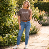 Woman wearing a brown t-shirt with 'five-oh' text, blue jeans, and white sneakers standing in a garden.