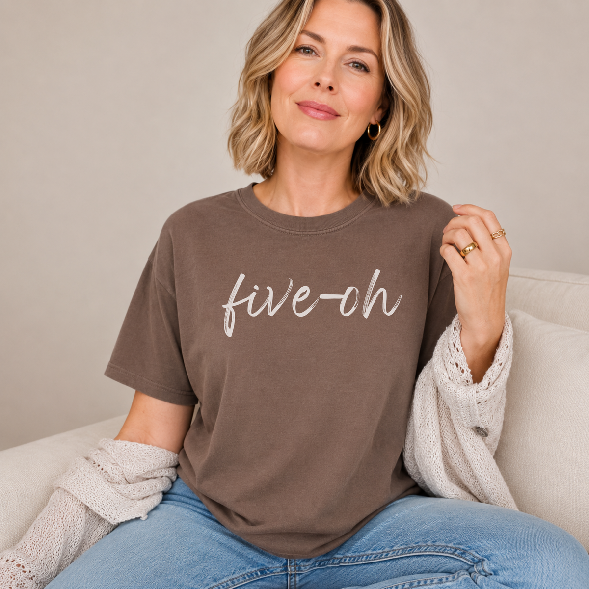 A woman sitting indoors with a espresso brown colored Comfort Colors shirt on with the words "five oh" written in script font.