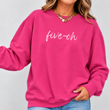 A woman wearing a bright pink crewneck sweatshirt with the text 'five-oh' written in white script across the chest, paired with blue jeans and a gold coin necklace, set against a plain light gray background.