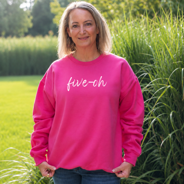 A woman wearing a bright pink crewneck sweatshirt with 'five-oh' written in white script across the chest, standing outdoors in a grassy park setting.