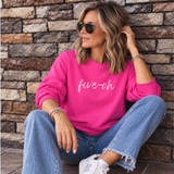 A woman wearing a bright pink crewneck sweatshirt with 'five-oh' printed in white script, paired with blue jeans and white sneakers, sitting against a rustic stone wall.
